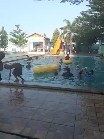 The video shows a group of children enjoying a swimming pool on a sunny day. Several kids are in the water, some using inflatable rings and boards. A yellow water slide is visible in the background. Two boys walk along the tiled edge of the pool. The surroundings include trees, a small building, and a tall structure.
