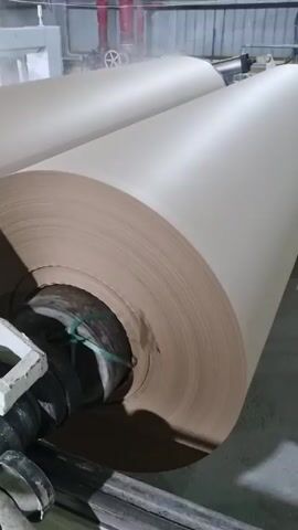 The camera shows a very large roll of kraft paper inside an industrial facility. There is a second large roll of the same paper behind the first. The paper is a light beige color and there are red pipes running across the ceiling. Some steam or fog is visible in the air.