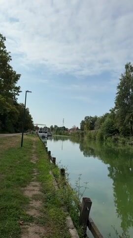 The video shows a serene canal scene with calm green water reflecting the sky and trees. On the left bank, a dirt path runs alongside the canal, bordered by grass and a low wooden fence with posts. A lamppost stands near the beginning of the path. Further down the left bank, several white boats are docked. The right bank is lined with lush green trees of varying sizes, creating a dense canopy. The sky is partly cloudy with patches of blue. The camera pans slowly to the right, revealing more of the tree line and the water's reflection.