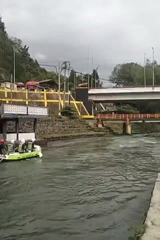 A small group of people are white-water rafting down a river in a green and yellow raft. They are all wearing life vests and helmets. The river is rough, with white water visible in the background. On the left bank of the river, there are some buildings with red roofs and yellow trim, as well as a hillside covered in trees. A stone staircase leads up from the river to a walkway with a white patterned railing. A large concrete bridge spans the river in the background, with an orange railing. The sky is overcast, suggesting a cloudy or potentially rainy day. The scene appears to be set in a semi-urban or tourist area, given the presence of buildings and infrastructure. The people in the raft appear to be enjoying the activity, though their specific emotions are not clearly discernible from their facial expressions. The overall mood is adventurous and active.