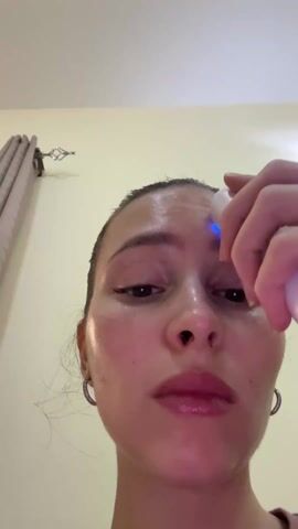 A woman with her hair pulled back is shown from a low angle, using a white and silver facial massager on her face. She starts on her forehead, then moves to her cheek and jawline. The device emits a blue light. The background is a plain wall with a curtain partially visible on the left side of the frame. The lighting is bright. The woman wears hoop earrings.