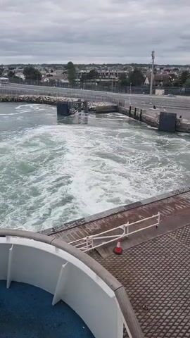 A video captures a wide shot of a river from a ferry. The water is churning, creating white foam. A concrete barrier is visible on the right, with a red cone. On the left, a white railing is visible. The background shows buildings, trees, a light pole, and a cloudy sky.