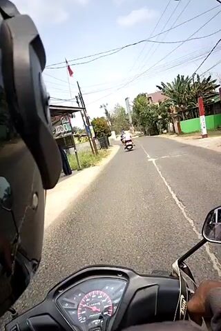 The video shows a first-person perspective of someone riding a motorcycle on a paved road. In the distance, several other motorcycles are visible, some with riders. The road is lined with trees, houses, and power lines. The sky is clear and the sun is shining. The rider's hands and part of the motorcycle's dashboard are visible in the foreground. The rider is following another motorcycle, which is also moving away from the camera.