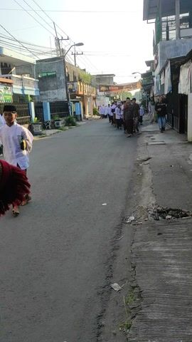 A group of people walks down a narrow residential street in a sunny urban setting. The group includes women in white head coverings, men in brown and white shirts and pants, and individuals in casual attire. Buildings line both sides of the street, showing a mix of residential and commercial structures. In the foreground, the corner of a red-patterned cloth and green clothing are partially visible, implying that the footage is taken from someone following or observing the group.
