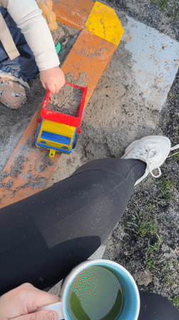 Child plays with toy truck in sandbox while adult watches
