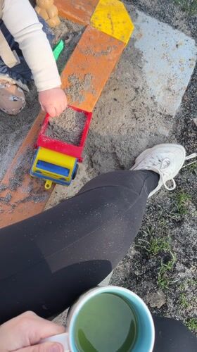 Child plays with toy truck in sandbox while adult watches