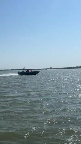 A motorboat with at least four people on board travels across a body of water under a clear sky. The boat is dark in color and appears to be moving at a moderate speed, as indicated by the spray of water behind it. The people in the boat are wearing life vests. The water is choppy, and the sun is reflecting off the surface. A shoreline with trees and vegetation is visible in the distance. A bird is flying in the sky above the boat.