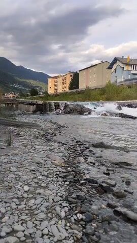 The video shows a river flowing over rocks, with a small waterfall in the middle ground. On the far bank of the river are several buildings, including apartment buildings and houses, situated on a slope leading up to mountains under a cloudy sky. The foreground is a rocky riverbed with scattered stones and pebbles. The water is clear and appears to be flowing moderately. No people are visible in the video. The overall scene depicts a natural landscape adjacent to a populated area.