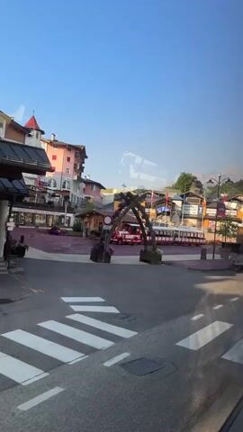 The video shows a street scene in a town, likely in the mountains. The buildings are a mix of architectural styles, with some having colorful facades. There is a small train with red and white carriages parked on a platform. The street is paved with asphalt and has white crosswalk markings. There are lampposts, flower pots with pink and purple flowers, and trees lining the street. In the background, mountains can be seen under a clear blue sky. Several cars are visible on the street, including a black car, a white van, and what appears to be a red fire truck on the train. The scene is well-lit, suggesting it is daytime. There are no people clearly visible in the video. The overall impression is of a quaint and picturesque town.