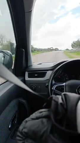 The video shows a car driving on a wet road. The view is from the driver's seat, with the seatbelt visible. In the distance, a white truck is driving on the same road, approaching the car. The weather appears to be overcast with patches of blue sky.