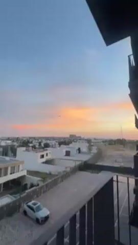 The video shows a residential area during sunset. The sky is a mix of blue, orange, and pink hues, indicating either sunrise or sunset. From a balcony, the view overlooks multiple white-roofed houses, a paved driveway with a silver car, and a fenced-off area. Solar panels are visible on the building's exterior. In the distance, there's an expanse of land, and a few taller buildings are present. A helicopter flies into view, silhouetted against the colorful sky. The video zooms in to showcase the helicopter more clearly before it flies out of frame.