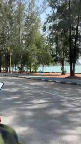 The video shows a view from a moving vehicle on an asphalt road. The road runs parallel to a waterfront. A row of tall, thin trees with green foliage lines the edge between the road and the waterfront. The water is a light turquoise color, and some boats are visible in the distance. A narrow pedestrian walkway, painted with a blue stripe, separates the trees from the road. Various vehicles, including a red ATV, a white pickup truck, a white sedan, and a motorcycle, are parked along the road. There's a white tent structure set up near the waterfront. The weather appears sunny with a clear blue sky. Hills or mountains can be seen in the distant background. The wind noise is significant throughout the video.