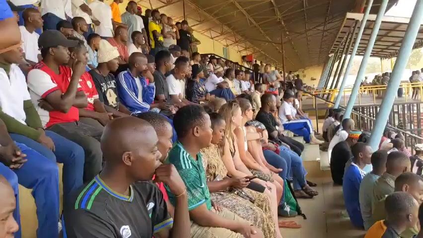 The video shows a crowd of people sitting in the bleachers of an outdoor stadium. Most appear to be watching a game or event taking place outside of the frame. The bleachers are partially covered by a metal roof structure. Many of the spectators are wearing casual clothing, including jeans, t-shirts, and hats. There is a mix of men and women of different ages and ethnicities. Some people are looking at their phones. Several are wearing athletic attire. The overall atmosphere seems relaxed and expectant. In the background, there are more people standing and walking around, and some trees are visible.