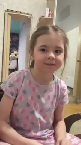 A young girl with missing front teeth laughs heartily and falls face-first into a brown sofa. She is wearing a light purple t-shirt with colorful heart shapes and light pink pants. Her brown hair is partially braided. The video is shot from indoors and shows a glimpse of another room with a chair, toys, and closet behind her. The wallpaper has a gray and white floral pattern.