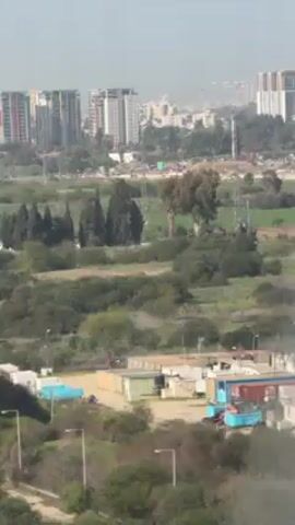 The video shows a view from an airplane window during takeoff or landing. Initially, the shot depicts a landscape with trees, fields, and tall buildings in the distance. As the plane moves, it passes by a group of parked airplanes, including a Boeing 747 with a distinctive red design near the front. The view then shifts to an area with airport buildings, open space, and a green field in the background. A security fence with a watchtower is also visible. Overall, the scene shows a mix of urban and natural environments, with a focus on the airport and its surroundings.