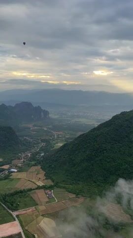 An aerial view of a valley with rolling green hills and cultivated fields. A hot air balloon is visible in the upper left corner of the frame. The sky is partly cloudy with rays of sunlight breaking through. The video captures the landscape from a high vantage point, offering a panoramic perspective of the natural environment. The clouds are gathering and moving across the sky. The sun's rays are diffused through the clouds, creating a serene and atmospheric scene. The valley below is dotted with small buildings and roads, suggesting a rural or semi-urban settlement. The overall impression is one of peaceful natural beauty and expansive scenery.