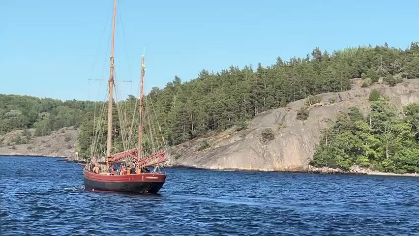 A wooden sailboat with red trim and two masts sails on blue water. The boat has a group of people on its deck. The boat is moving from left to right across the frame, creating a wake. In the background, a rocky, tree-covered island rises from the water under a clear blue sky. The sun is shining, and the water is calm with small waves.