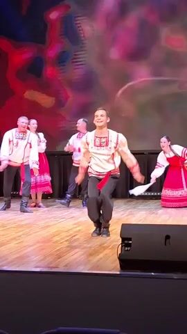 A group of dancers in traditional Russian attire perform on a wooden stage. The lead dancer, a man in a white embroidered shirt with red trim and dark pants, is captured mid-air in a high jump with his legs spread wide. Several other male and female dancers in similar colorful outfits are also on stage, some clapping or holding white sashes. The background features a projection of a colorful abstract design, which transitions to a landscape scene with rolling green hills. The stage is lit by spotlights, and black speakers are visible in the foreground.