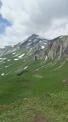 A person is panning across a vast mountain landscape under a partly cloudy sky. The terrain is characterized by steep, green slopes dotted with patches of snow. The foreground consists of a grassy slope with scattered wildflowers. In the distance, several layers of mountains are visible, some covered in dense forest and others with lingering snow. The overall impression is one of rugged, natural beauty in a high-altitude environment.