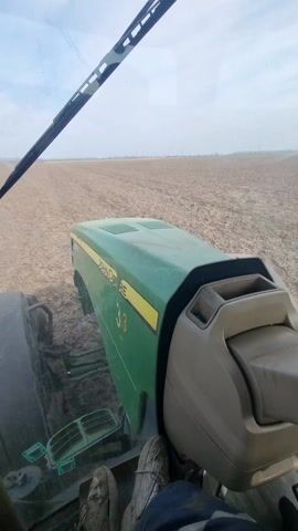 The video, filmed from inside the cab of a green John Deere tractor with the number 33 on the hood, shows the driver operating the tractor in a dry, plowed field. The view is through the front windshield, showing the vast expanse of the field, and briefly through the side mirror, revealing a water tank being towed behind the tractor. The sky is mostly clear with a few wispy clouds. The driver's feet, clad in sneakers and jeans, are visible in the foreground. Music is playing.