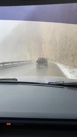 The video, filmed from inside a car, shows a first-person perspective of driving on a wet, winding mountain road in what appears to be snowy or misty conditions.

In the foreground, the dashboard and windshield of the recording vehicle are visible. An orange light displaying "ON" and "PASSENGER AIRBAG" is illuminated on the dashboard. The wipers are likely in operation, given the wet windshield, although their movement is not clearly captured.

Ahead, a dark-colored SUV or pickup truck is being followed. This vehicle has items, possibly camping gear or equipment, secured in its open cargo bed. Further down the road, another smaller car can be seen ahead of the SUV. The road itself is asphalt, appearing wet, and traces of snow or ice are visible on the shoulders and adjacent terrain. To the right, a steep, rocky mountain face dominates the view, partially obscured by mist or fog, which gives the scene a hazy, diffused light. To the left, a metal guardrail follows the curve of the road.

The overall scene depicts cautious driving through a mountainous region, characteristic of areas like Lago Escondido, Argentina, which is known for its rugged terrain and variable weather. The conditions suggest cold temperatures and reduced visibility, requiring careful navigation.