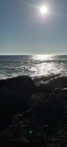The video shows a view of the ocean from a rocky shore. The sun is shining brightly, creating a glare on the water. Waves are crashing against the rocks. The rocks in the foreground are dark and appear wet. The sky is clear and blue.
