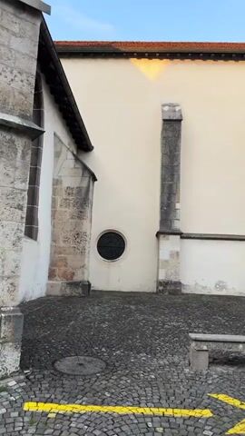 A vertical video pans across the exterior of a stone building, likely a church, with cobblestone paving in the foreground. On the left, a section of a stone tower with a pointed arch is visible. To the right of the tower, a pale wall is seen, punctuated by a small, circular, dark-colored window with a decorative grate. Further to the right, a tall, narrow stone buttress extends upwards. The building has a tiled roof, and sunlight casts a triangular reflection on the pale wall. A planter with a dry-looking shrub is positioned near the base of the wall. Towards the end of the video, the camera moves to reveal a large wooden double door with decorative metalwork, approached by stone steps. A black and white striped pole with a metal railing stands to the right of the entrance. A drain grate is visible on the cobblestones in the lower right corner. The sky above is a clear blue. No people or animals are present.
