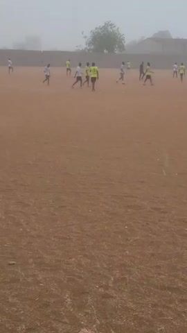 The video shows a soccer match in progress on a dirt field during a foggy or hazy day.  Several players wearing light-colored shirts and some wearing bright yellow-green shirts are scattered around the field, moving and kicking a ball. There is a goalpost visible at one end of the field. A large tree stands in the middle of the field, and a wall borders the playing area in the background.  The ground is dry and brown. The overall visibility is low due to the fog.