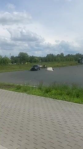 A black Subaru Impreza is shown driving on a paved track, possibly a race track or a driving course. The car appears to be drifting or performing maneuvers. The track is surrounded by green grass and trees. In the background, there are some buildings, a light pole, and other cars. The sky is cloudy. The video is shot from a slightly elevated position, looking down at the car. There are some orange cones and a white barrier on the track. The car has a red sticker on its windshield.