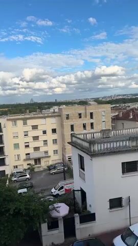 The video presents a static, high-angle view of a residential area on a partly cloudy day. Several apartment buildings of varying heights and colors dominate the foreground. Cars are parked on the street and in a small parking lot visible between buildings. Green trees are interspersed among the buildings. In the distance, a cityscape is visible, with the Eiffel Tower and another tall building distinguishable on the horizon. The sky above is blue with scattered, puffy white clouds. The overall scene suggests a suburban or urban residential area on a pleasant day.