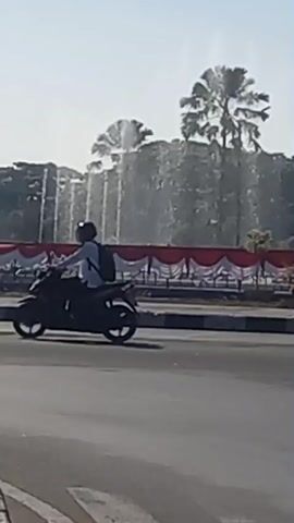 The video shows a road with a roundabout in the background. A fountain is active in the roundabout, with water spraying upwards. The roundabout is decorated with red and white fabric. Palm trees are visible behind the fountain. A black Suzuki Ertiga car drives past the fountain. Several motorcyclists are also seen riding on the road. The sky is clear and the sun is shining.