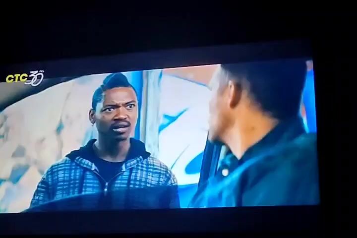 The video shows a screen displaying a scene from a movie or TV show. In the foreground, on the left side of the screen, is a man of African descent with a distinctive hairstyle. He is wearing a plaid hooded jacket over a dark shirt and appears to be speaking emphatically. He is looking towards the right of the screen. On the right side of the screen, partially obscured, is another person, likely male, seen from the back of his head and shoulders, wearing a dark jacket. They appear to be in a car, indicated by the car door frame visible in the foreground and the presence of glass reflections. The background is out of focus and appears to be an urban or industrial setting with graffiti or painted walls. A channel logo "CTC 30" is visible in the top left corner. The scene appears to be a tense conversation or confrontation.