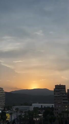 A time-lapse video shows a sunset over a mountainous landscape, with buildings and palm trees in the foreground. The sky transitions from a pale blue to a warm orange as the sun dips below the horizon. The video captures the changing light and colors of the sky.