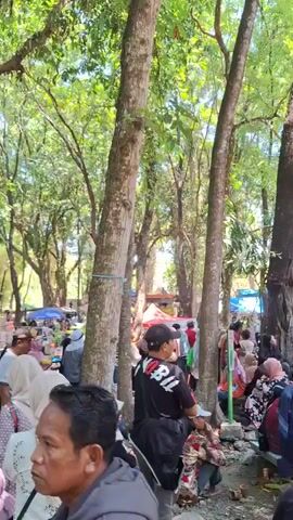 The video shows a crowd of people gathered under large trees with lush green foliage. The camera pans from the foreground where several individuals are seated on the ground and on a bench, to the middle ground where more people are standing and interacting. In the background, there appear to be market stalls and some sort of structure. The lighting suggests it is daytime, and the environment is outdoors. The people are dressed in casual clothing, some wearing headscarves. The overall impression is of a lively gathering or event in a shaded, natural setting.