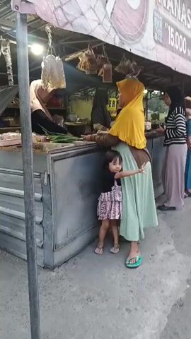 A group of women and a young child are standing at a market stall. One woman, wearing a yellow hijab and a green dress, is holding a small child who is wearing a pink dress. Another woman, wearing a black and white striped top and a pink skirt, is standing behind them. A third woman, wearing a blue dress, is standing further down the stall. The stall appears to be selling fruit, with a sign that says "NANAS 15,000." There are motorcycles parked on the side of the road in the background. The ground is paved and there is some debris on it. The lighting suggests it is late afternoon or early evening.