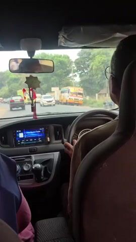 The video begins inside a vehicle, looking forward through the windshield. A person wearing glasses is driving, with their hands on the steering wheel. A person in a dark blue hijab is seated in the passenger seat, partially visible. The dashboard features a touchscreen infotainment system, air vents, and a gear stick. A decorative item with red tassels hangs from the rearview mirror. Outside, the road is busy with various vehicles, including cars, motorcycles, a white SUV, and a large green dump truck. The surroundings appear to be a semi-urban or rural area with trees and utility poles visible along the roadside. The weather seems overcast. The camera pans slightly as the vehicle moves forward.