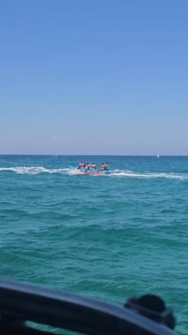 A bright, clear day with a blue sky and blue ocean. Several people are riding on a banana boat, which is being pulled through the water by a boat that is out of frame. The banana boat is colorful, with blue and pink accents. The people on the banana boat are wearing swimsuits and appear to be enjoying themselves. The water is choppy, with whitecaps visible. The overall scene is one of summer fun and recreation.
