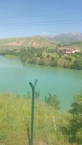 The video shows a scenic view of a lake with turquoise-colored water, captured from a moving vehicle, possibly a train. Lush green vegetation, including trees and bushes, lines the lake's edge in the foreground. In the background, rolling hills and distant mountains with patches of snow on their peaks are visible. A few buildings are scattered across the landscape, along with power lines stretching across the sky. The sky is clear and blue, indicating a sunny day. The camera moves at a steady pace, offering a continuous view of the landscape as the vehicle progresses.