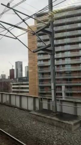 The video shows a view from a moving train, looking out towards a city skyline. The foreground features a grey concrete wall, railway tracks, and overhead power lines.  In the midground, there are several buildings of varying sizes and styles, including a modern high-rise with a facade of stacked, golden-colored rectangles. In the background, a cluster of taller, glass-covered skyscrapers dominates the horizon.  The sky is overcast and grey.  The train is moving at a moderate speed, causing the buildings to gradually pass by.