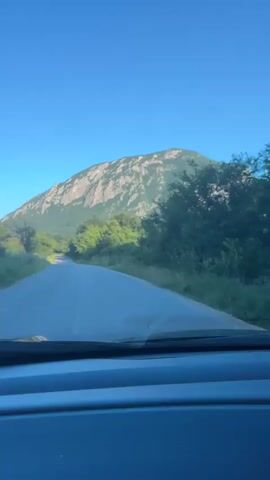 A car drives along a paved road towards a prominent, tree-covered mountain under a clear blue sky. The road is flanked by green grass and lush trees. The mountain, with some visible rocky outcrops, dominates the horizon. The scene appears to be set on a bright day, possibly morning or late afternoon, given the clear conditions and lighting. The view is captured from inside the moving vehicle, with the dashboard visible at the bottom of the frame. This appears to be a scenic drive in Glavatsi, Bulgaria.