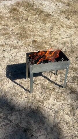 The video captures a serene outdoor scene on a sunny day at a beach in Zīvartiņš, Latvia. The primary subject is a metallic brazier, commonly known as a mangalas, placed on the sandy ground. Inside the brazier, a fire is actively burning, with visible flames and glowing embers, suggesting that wood or charcoal is being prepared for grilling or barbecuing.

The ground is composed of light-colored sand, characteristic of a coastal area, and features sparse patches of dry, light-brown grass, indicative of beach dunes. As the camera slowly pans upwards, a wide expanse of the Baltic Sea becomes visible in the background, with gentle waves lapping at the shore. The sky above is a clear, vibrant blue with minimal wispy white clouds, reflecting bright daylight. The strong sunlight casts a distinct, dark shadow of the brazier on the sand.

No people are visible in the footage, emphasizing the natural beauty of the seaside environment and the simple act of preparing a fire outdoors. The scene conveys a sense of calm and anticipation for an outdoor meal by the sea. There is no discernible text or branding on any objects within the video.