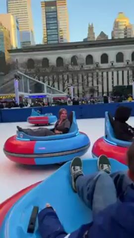 The video shows people riding bumper cars on an ice rink. The bumper cars are primarily blue and red, with a visible number on the front. Riders are seen sitting in the cars, navigating around the ice. In the background, tall buildings are visible under a bright sky, along with a large building behind the ice rink. The video appears to be shot at ground level, following the motion of one of the bumper cars.