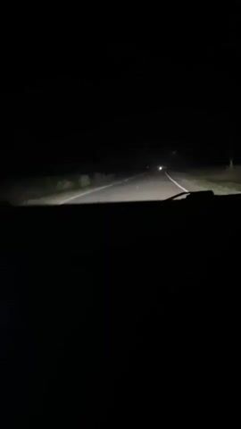 The video shows a first-person perspective from inside a vehicle driving on a dark, paved road at night. The headlights illuminate the road ahead, revealing white lane markings and the edges of the road, which appear to be bordered by vegetation. In the distance, there is a bright light, possibly from another vehicle's headlights. The interior of the car is dark, with only a portion of the dashboard visible. The overall scene conveys a sense of nighttime travel through a rural or unlit area.
