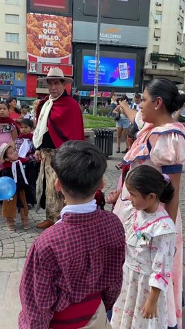 A group of Argentinians, adults and children, stand on a cobblestone square in a city, dressed in traditional clothing. One child holds a blue and white Argentinian flag, while another holds a blue balloon. The adults wear hats and capes, and the children wear dresses with floral patterns or traditional pants and vests. Several buildings are visible in the background, featuring large advertisements for KFC, Burger King, and other businesses. People walking through the square watch the group.