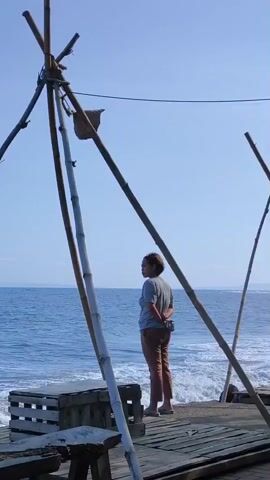 A woman with short brown hair, wearing a light-colored t-shirt and reddish-brown pants, stands on a wooden pier or platform, looking out at the blue ocean. Her back is partially to the camera, and her hands are clasped behind her. To her right, a tall, crude tripod-like structure made of bamboo poles stands upright, with a small woven bag hanging from one of the poles. Another similar structure is partially visible further right. In the foreground to her left, a wooden crate is visible. The sky is clear and blue, and the ocean has gentle waves breaking near the platform. The scene is brightly lit by the sun.