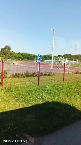 The video shows a bright, sunny outdoor scene. In the foreground, there is green grass with some small flowers and the shadow of a person or object. A metal fence with red posts separates the grassy area from a red and grey athletic track. Beyond the track, there is a tower with a ladder, a light pole, and what appears to be a parking area with a blue sign indicating parking. Trees and more athletic field equipment are visible in the background under a clear blue sky. The bottom left corner of the video displays "Infinix HOT 50." The timestamp at the beginning reads "00:00."