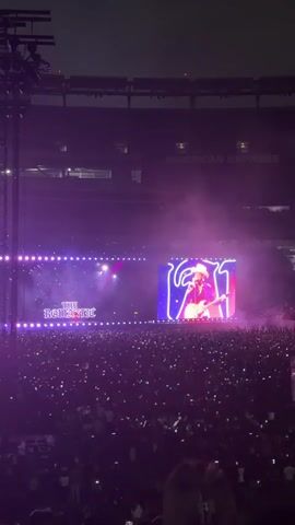 The video shows a large outdoor concert at night. A massive crowd of people is visible in the foreground, with many holding up their phones, creating a sea of lights. On a stage in the background, a large screen displays a performer wearing a cowboy hat and playing a guitar. The stage is lit with vibrant purple lights and smoke effects. Another screen on the left side of the stage displays text that reads "THE ROMANTIC". The overall atmosphere appears energetic and festive.