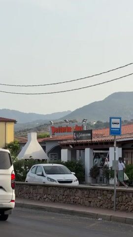 This video captures a street view with a building in the background that appears to be a market or restaurant, indicated by the sign "Detturmarket" and another sign that says "La Gallura Gelateria Restaurant." A white car is parked in front of the building, and people are visible near the entrance of the establishment. To the right of the building, there is a blue bus stop sign. In the foreground, a gray car drives past, partially obscuring the view of the building and the street. The background shows hills or mountains under a clear sky. The scene appears to be an outdoor commercial area with moderate traffic.