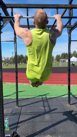 A man with red hair, wearing a yellow tank top, yellow shorts, and yellow sneakers, performs a pull-up on a pull-up bar located on a platform outside on a sunny day. The man has a tattoo on his upper left arm. A red running track and sports field can be seen in the background with people playing. There are some trees, tents, and a water bottle visible in the background as well. The pull-up bar structure is made of black metal with the word "VIVUSTEEL" on it. The man does one pull-up, releases the bar, turns, and walks away. He's wearing sunglasses.