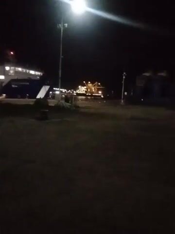 The video shows a nighttime scene at a dock. A ferryboat is visible on the left, illuminated by lights. In the background, another larger vessel or structure, possibly an oil rig, is also lit.  A street light shines brightly on the left side of the frame. The ground is dark, appearing to be pavement or a similar surface. There are no people visible.  The camera appears to be stationary, and the video is oriented vertically.